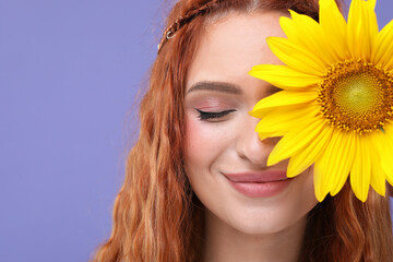 Beautiful young hippie woman covering eye with sunflower on violet background, closeup