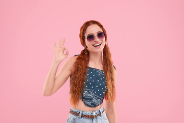 Stylish young hippie woman in sunglasses showing OK gesture on pink background