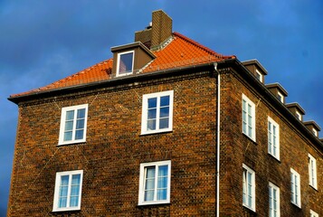 Altes rotbraunes Backsteinhaus mit weißen Fensterrahmen und rotem Ziegeldach vor blauem Himmel in Stadt bei Sonne und Regen am Nachmittag im Herbst