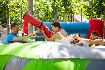 Fototapeta na wymiar Adults having fun on inflatable amusement playground. Emotional young girl fighting off her friends with inflatable log while they trying to filch toy chickens