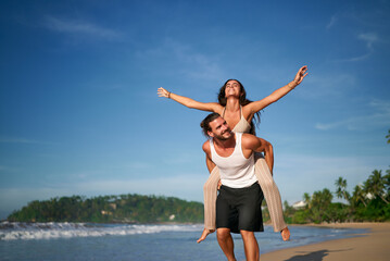 Joyful man carries girlfriend on his back, woman raises arms embracing freedom on tropical beach....