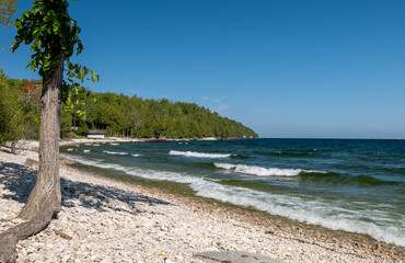 White waves roll onto a rocky tree-lined shore on a sunny day, with the horizon over the blue water in the distance.