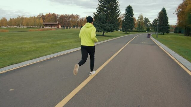 Back view of sporty man in black bini hat, tracksuit of bright color exercising in nice park with green lawns, evergreen trees and colorful autumn trees. High quality 4k footage