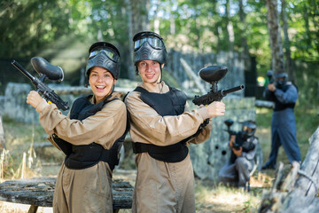 Portrait of positive young woman and man paintball players stanidng back-to-back on battlefield.