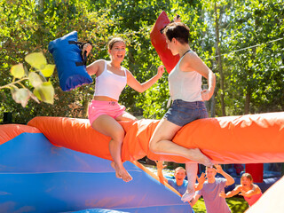 Laughing young girl fighting by pillows with female opponent while sitting on inflatable beam on...
