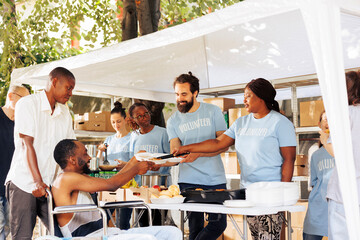 Free meals and fruits are given to a wheelchair-bound african american man in need. Smiling volunteers of different ethnicities aiding the handicapped poor and homeless people at outdoor food bank. - obrazy, fototapety, plakaty