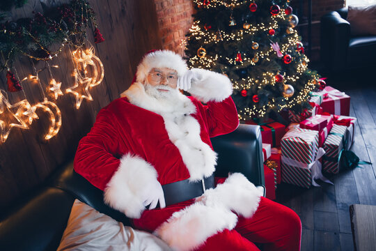 Photo of positive cheerful santa claus wear red costume spectacles sitting cozy sofa indoors new year north pole office