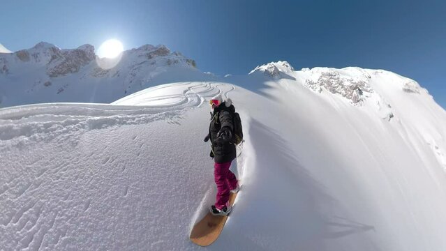 SELFIE, LENS FLARE: Female snowboarder riding beautiful snowy mountain terrains. Winter adrenaline enthusiast sprays clouds of fresh snow while snowboarding untracked terrain on a splitboarding trip.