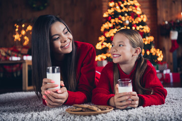 Photo of cute dreamy small sisters wear red jumpers lying floor drinking cacao eating chocolate biscuits indoors new year north pole house