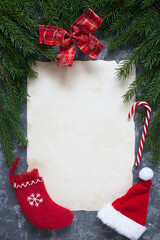 Christmas background with fir branches, red ribbon bow and old paper, stocking with a gift, lollipop, Santa hat on gray table. Copy space, top view.