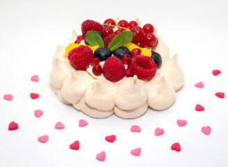 Meringue pavlova dessert, decorated with whipped cream,  berries and mango. Surrounded with heart shaped sprinkles. Photographed against a white background.