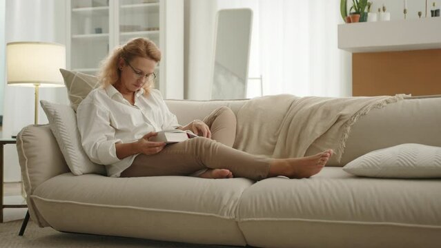 Tired lady in glasses with thick book in hands sitting with streched legs on sofa, closing eyes, benting head over the book. Light room with renovation. High quality 4k footage