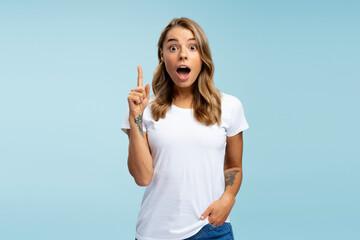 Funny emotional student holding finger up having creative idea isolated on blue background, education concept. Amazed attractive young woman with open mouth pointing hand looking at camera in studio