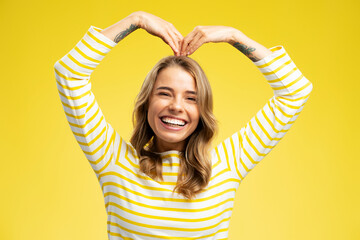 Smiling beautiful woman, cute overjoyed influencer with wavy silky hair wearing stylish striped sweater  showing heart shape isolated on yellow  background. Love, positive lifestyle concept