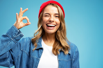 Smiling beautiful woman winking wearing stylish red hat showing ok  sign isolated on blue background. Portrait happy modern hipster female with white teeth looking at camera in studio, dental concept  - obrazy, fototapety, plakaty