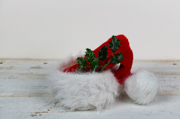 Red and white furry Santa Claus hat with a sprig of holly isolated on a wood plank table with a shallow depth of field and copy space