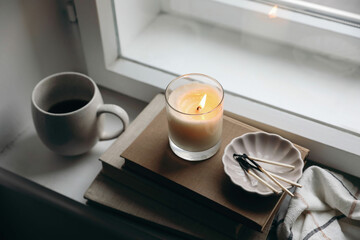 Moody autumn, winter still life. Burning candle on old books. Matches, ceramic plate on windowsill. Blurred cup of coffee, mug. Cozy fall mood. Thanksgiving. Halloween. Top view, selective focus.