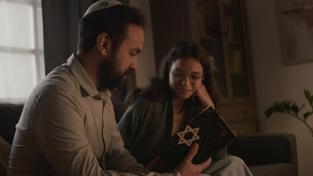 Medium side shot of Jewish dad in kippah and young daughter sitting together on couch at home, holding Torah book with star of David, reading prayers, studying and discussing scripture