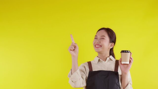 Portrait of young asian barista woman wearing apron holding coffee cup and presenting on yellow background, waitress or entrepreneur cheerful and showing, small business or startup, waiter of cafe.