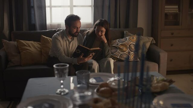 Full shot of bearded Jewish man in kippah sitting on couch with young girl, holding Torah book, reading and discussing holy scripture, and festive table with hanukkiah in blurred foreground