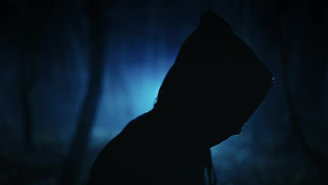 A dark figure in a cloak with a hood in a sinister night forest. Fantasy demon or witcher in the night.