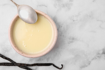 Bowl with condensed milk, vanilla pods and spoon on white marble table, flat lay. Space for text