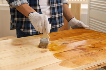 Man with brush applying wood stain onto wooden surface indoors, closeup