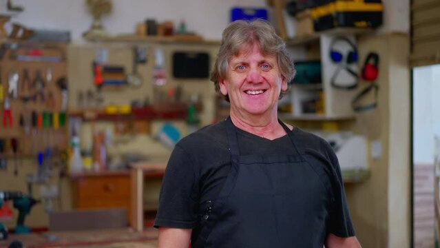 One joyful master carpenter portrait smiling at camera, wearing apron standing inside carpentry workshop with tools and equipment in background hanging on the wall
