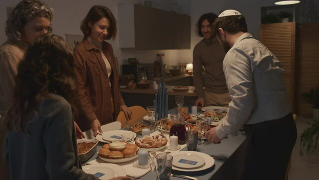 Medium shot of Jewish family of five, men wearing kippah, getting ready for Hanukkah dinner together at home, putting cutlery and food on table, chatting and smiling