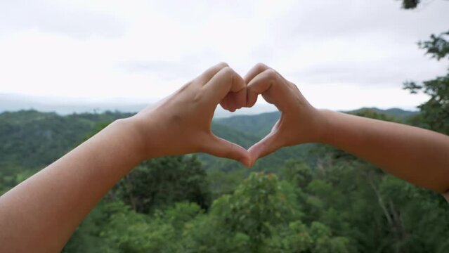 Two hands joined together to form a heart shape, a symbol of love and affection against a natural background of mountains and trees.
