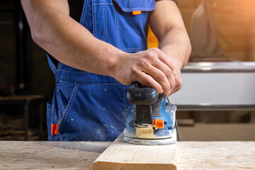 Young man builder carpenter equals polishes wooden board with a  random orbit sander  in the...