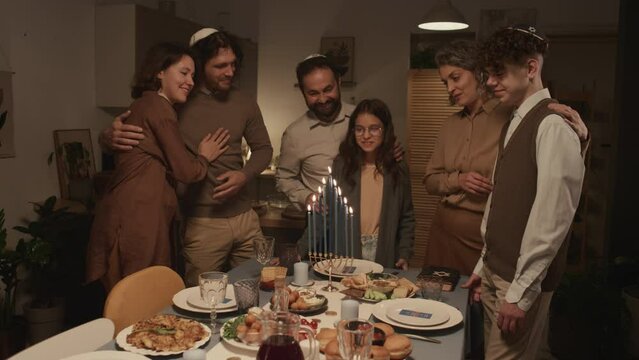 Full shot of happy Jewish family of six standing around festive table with traditional foods, looking at burning candles on menorah on final day of Hanukkah holiday, smiling and hugging