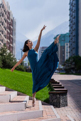 Fototapeta na wymiar Beautiful Asian ballerina in a blue dress stands on the stairs in the splits outdoors. Urban landscape. Vertical photo. 