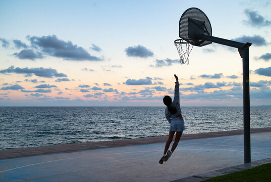 Silhouette of a girl jumping towards the basket, on a basketball court in front of the beach, at sunset.