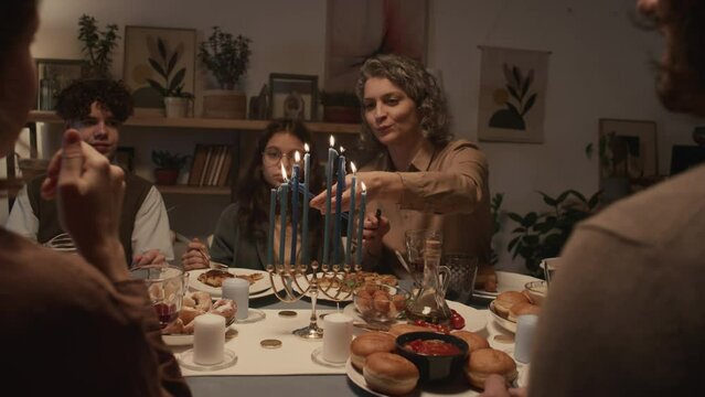 Medium over-shoulder shot of Jewish mother sitting at table with family during Hanukkah dinner, lighting up candles on hanukkiah as her son and daughter are watching, and chatting to guests