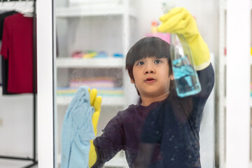 Kid little asian boy son having fun doing household cleaning and washing in protective gloves using a spray and rag while cleaning window in living room at home.housework and home cleaning concept