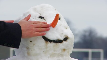 Detailed view of a man’s bare hands carefully placing a carrot nose and topping the snowman with an orange hat — a cozy moment of winter craftsmanship and festive spirit. - Powered by Adobe