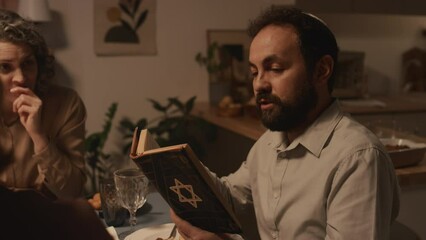 Medium shot of middle-aged Jewish man in kippah sitting at festive dinner with family, celebrating Hanukkah in candlelight, reading passages from holy Torah with star of David on cover - Powered by Adobe