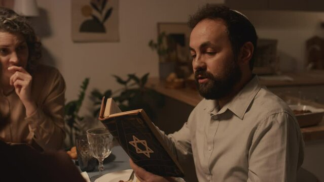 Medium shot of middle-aged Jewish man in kippah sitting at festive dinner with family, celebrating Hanukkah in candlelight, reading passages from holy Torah with star of David on cover