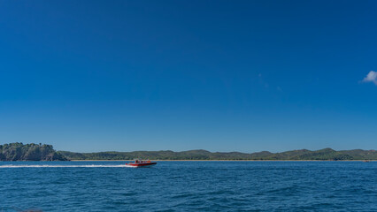 A boat with tourists is rushing across the blue ocean. Foam trail behind the stern. Green coastal hills against the azure clear sky in the distance. copy space. Madagascar.