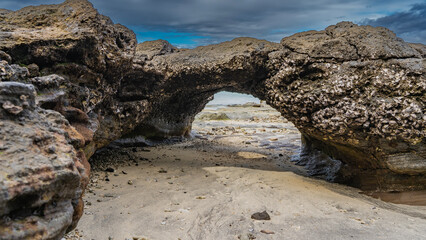 A bizarre stone grotto was exposed at low tide. Attached mollusks are visible. The ocean is visible through the arch. Clouds in the blue sky. Madagascar. Nosy Tanikeli   