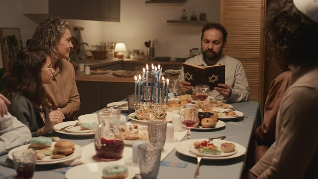 Medium full shot of middle-aged Jewish man in yarmulke sitting at Hanukkah feast with family, reading text from holy Torah with star of David on cover, and candles burning on hanukkiah on table
