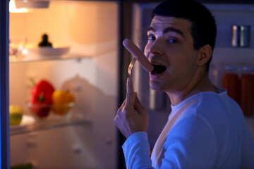 Hungry young man eating sausage near open fridge in kitchen at night, closeup