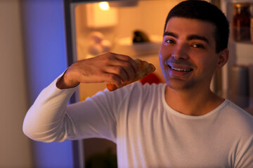 Hungry young man with croissant near open fridge in kitchen at night, closeup