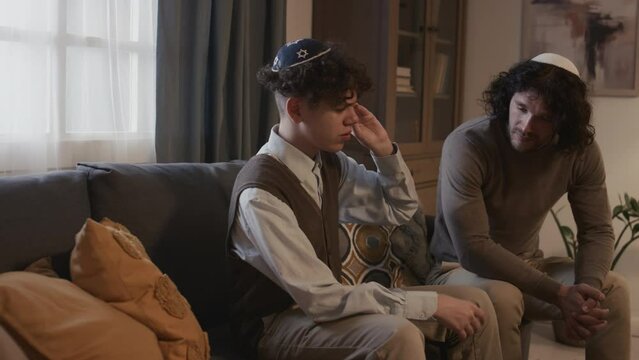 Medium shot of unhappy teenage Jewish boy with curly hair, in kippah sitting on couch with older brother during family gathering on Hanukkah, talking, sharing problems and asking for advice