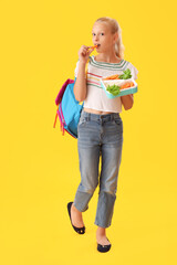 Girl with backpack and lunchbox eating vegetables on yellow background