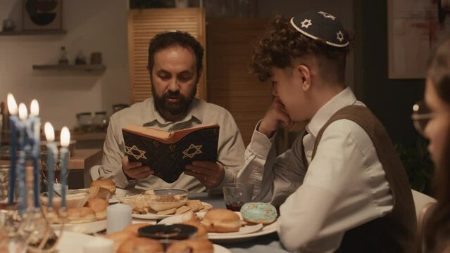 Medium shot of devoted Jewish father in kippah sitting at Hanukkah dinner with family and reading story from holy Torah to teenage son in skullcap and daughter, children listening attentively