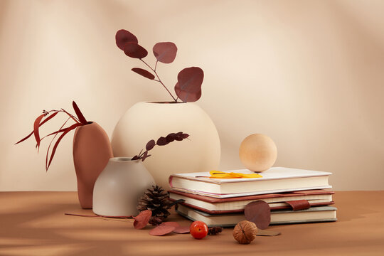 Stacked notebooks, autumn leaves, ceramic vases, and herbs on a brown table against a beige background. Front space adorned with a fall theme.