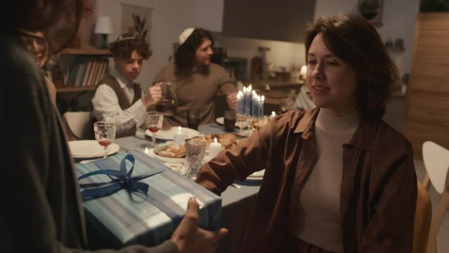 Medium shot of young Jewish woman giving gift to younger sister and wishing her good things at Hanukkah celebration, while family members are sitting at festive table and preparing to eat dinner
