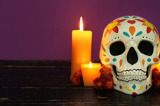 Painted human skull for Mexico's Day of the Dead (El Dia de Muertos) with burning candles and flowers on wooden table against purple background